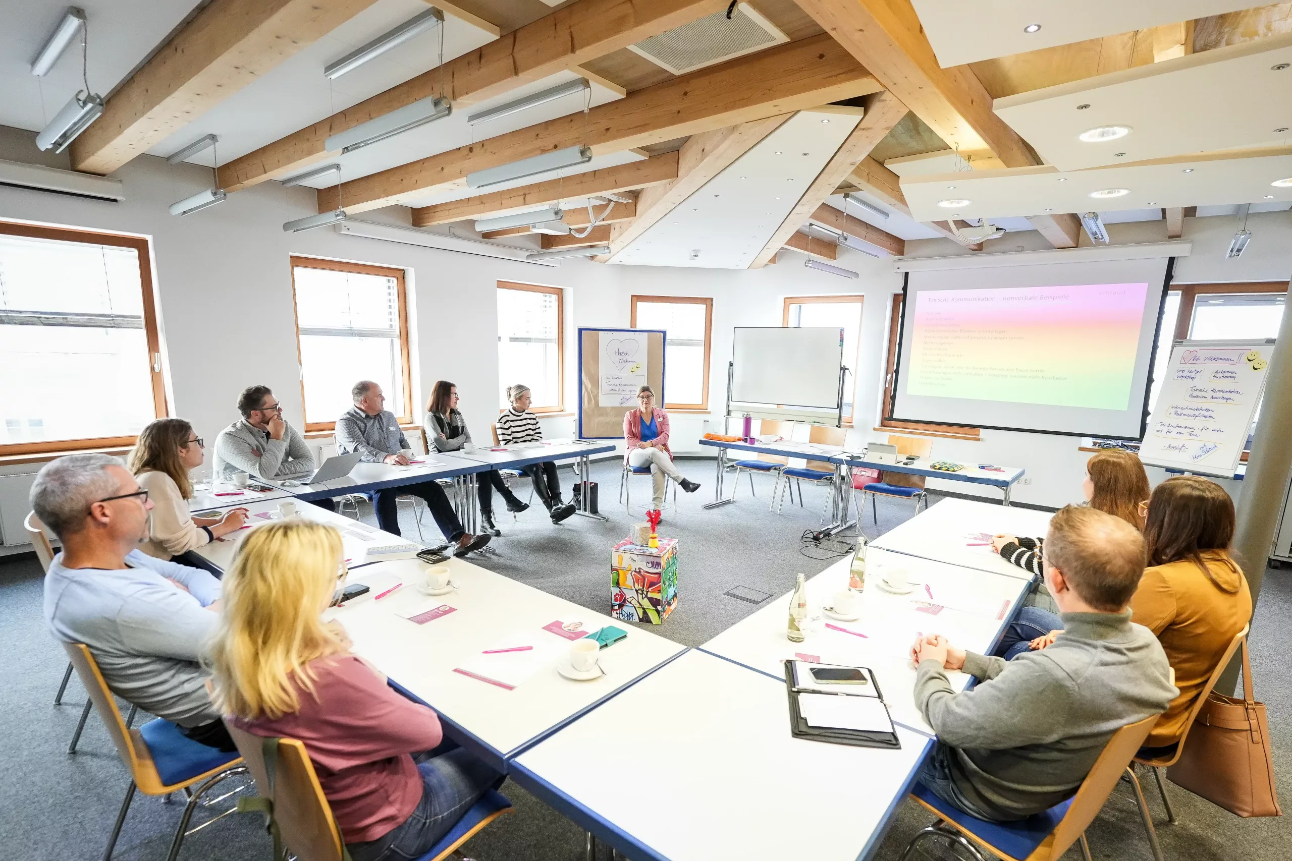 Eine Gruppe von Personen sitzt in einem hellen Besprechungsraum an U-förmig angeordneten Tischen und hört aufmerksam einem Vortragenden zu, der vor einem Bildschirm sitzt, auf dem eine Präsentation gezeigt wird.