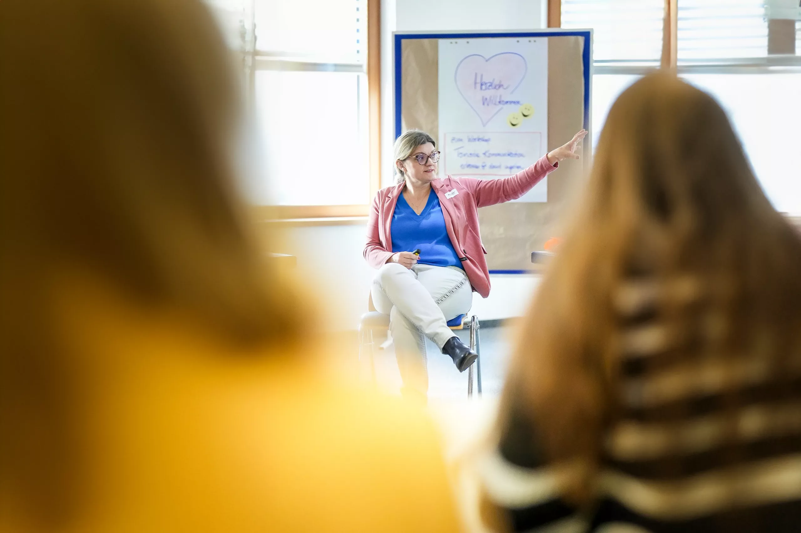 Eine Frau sitzt auf einem Stuhl und gestikuliert, während sie zu einer Gruppe spricht. Hinter ihr steht ein Flipchart mit einem Herz und dem Wort Willkommen. Im Vordergrund sind zwei verschwommene Personen zu sehen, die ihr zuhören.